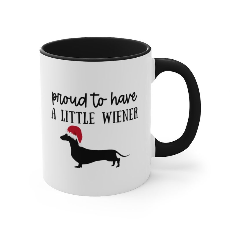 May include: White ceramic mug with a black interior and handle. The mug features the text "proud to have a LITTLE WIENER" above a black dachshund wearing a red Santa hat. A fun, festive gift for dog lovers.