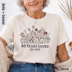 May include: A cream-colored short-sleeve t-shirt featuring a graphic of various wildflowers and a small butterfly. Below the floral design, the text reads 80 YEARS LOVED Est. 1946 in a dark gray font. The shirt has a crew neckline and a standard fit.