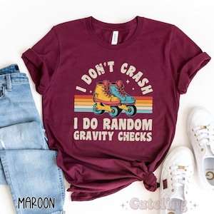 Pu&ograve; includere: Una maglietta color granata con un design retr&ograve; che presenta un paio di pattini a rotelle e il testo "I don't crash I do random gravity checks".