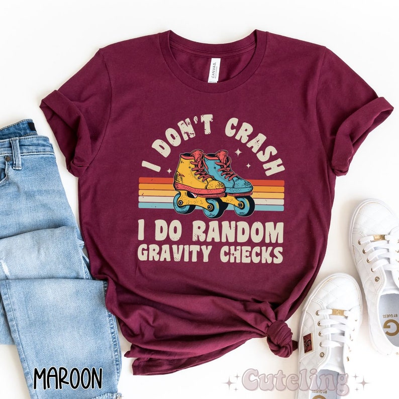 Pu&ograve; includere: Una maglietta color granata con un design retr&ograve; che presenta un paio di pattini a rotelle e il testo "I don't crash I do random gravity checks".