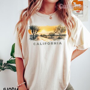 May include: A cream colored t-shirt with a watercolor illustration of a California sunset with palm trees and the word "CALIFORNIA" printed below the image. The t-shirt is labeled "IVORY".