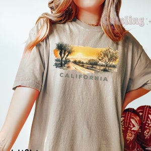 May include: Khaki t-shirt with a watercolor illustration of a California desert scene with palm trees, a road, and the word "CALIFORNIA" printed below the image.