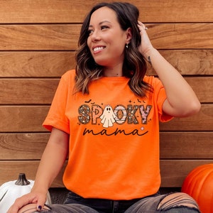 May include: A woman wearing an orange t-shirt with a ghost and the text "Spooky mama" printed on it. She is standing in front of a wood panel background.