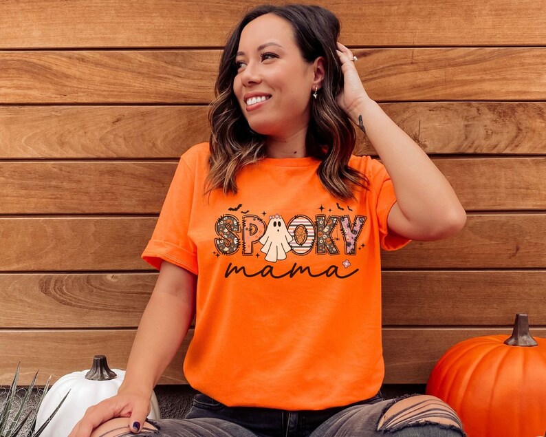 May include: A woman wearing an orange t-shirt with a ghost and the text "Spooky mama" printed on it. She is standing in front of a wood panel background.