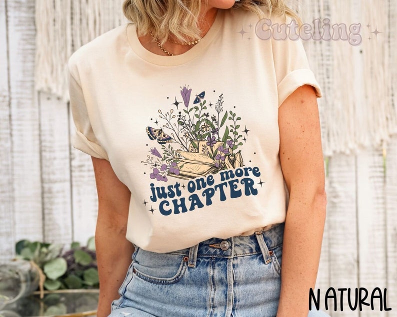Puede incluir: Una camiseta de color crema con un gr&aacute;fico de una pila de libros con flores y mariposas. El texto "Just one more chapter" est&aacute; escrito en azul en la camiseta.