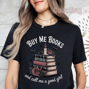 May include: Black t-shirt with the text "BUY ME BOOKS and call me a good girl." The graphic features a stack of books, roses, butterflies, and a candle with a lit flame. The design is in white, pink, and black.