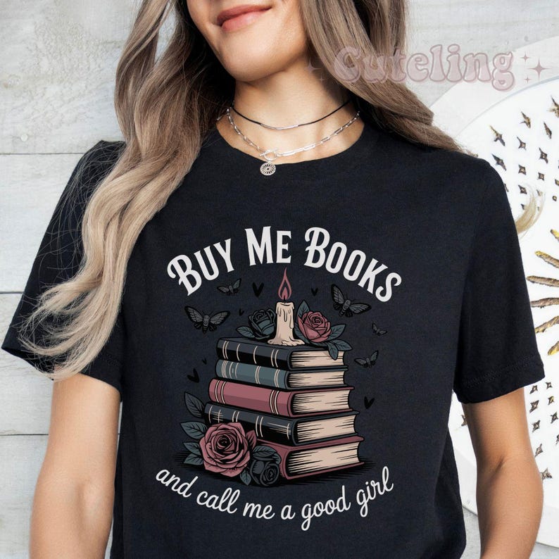 May include: Black t-shirt with the text "BUY ME BOOKS and call me a good girl." The graphic features a stack of books, roses, butterflies, and a candle with a lit flame. The design is in white, pink, and black.