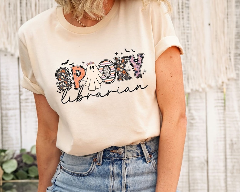 May include: A cream-colored t-shirt with a graphic design of a ghost and the text "Spooky Librarian" in colorful patchwork letters.
