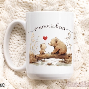 Peut inclure: Mug en céramique blanche avec un ours brun et un ourson brun sur un pont de bois. Le plus grand ours tient un ballon rouge en forme de cœur. Le texte "mama bear" est écrit en cursive sur la tasse.