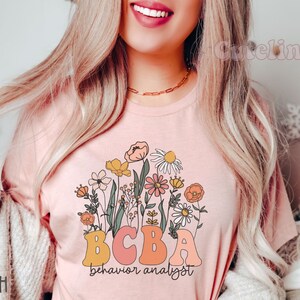 Pu&ograve; includere: Una maglietta rosa con un design floreale e il testo "BCBA behavior analyst".