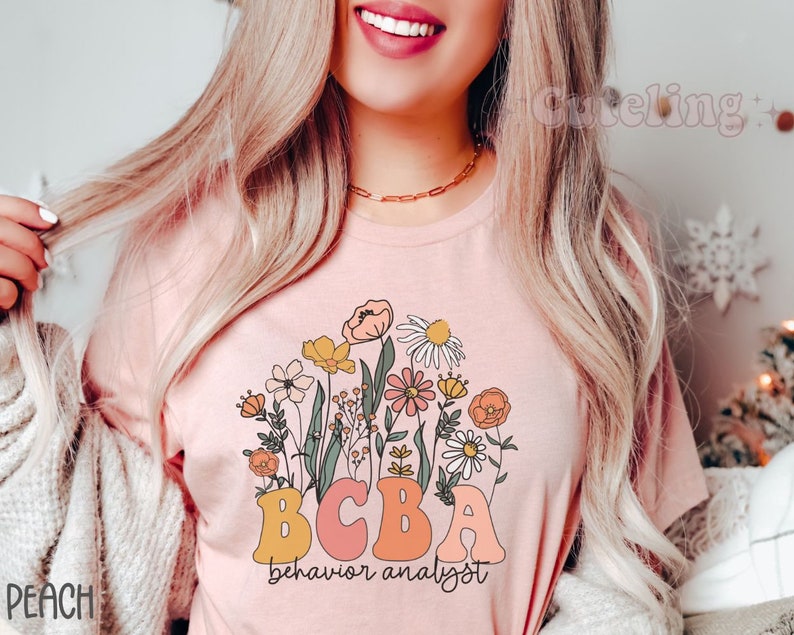 Pu&ograve; includere: Una maglietta rosa con un design floreale e il testo "BCBA behavior analyst".