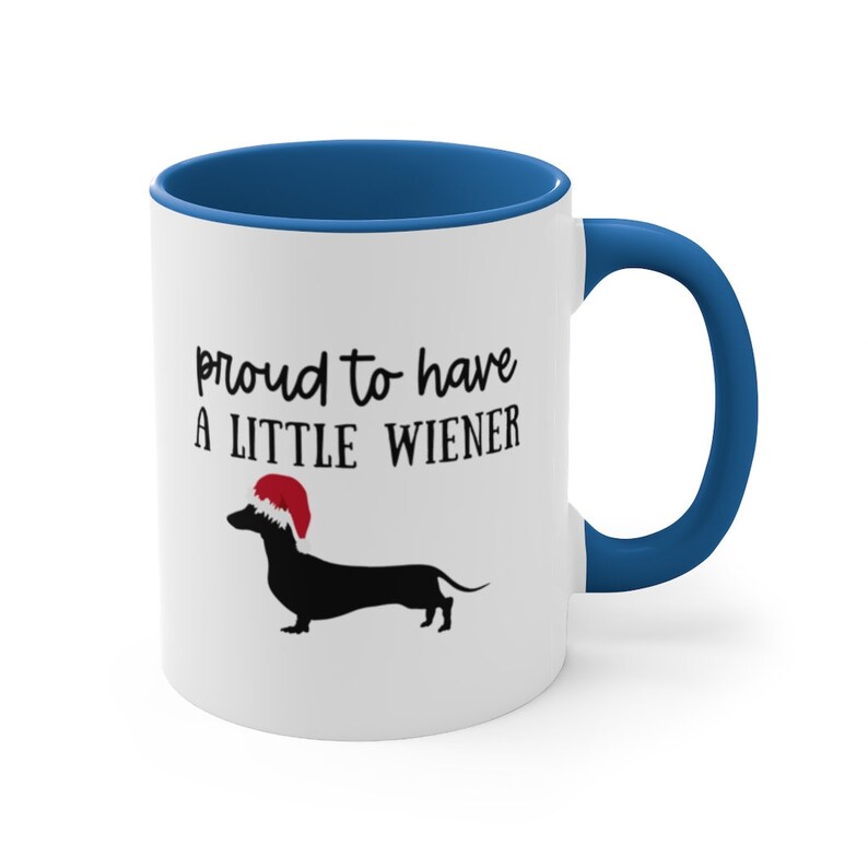 May include: White ceramic mug with a blue interior and handle. The mug features the text "proud to have A LITTLE WIENER" with a black dachshund wearing a Santa hat.