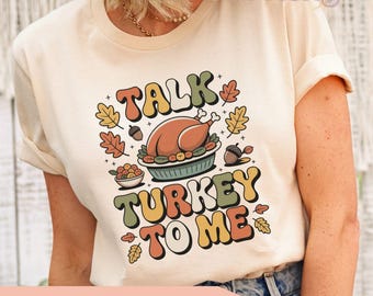 May include: A cream-coloured t-shirt with a retro graphic that says "Talk Turkey To Me." The design features a roasted turkey, autumn leaves, and acorns. The text "More Colors Available" is printed below the graphic.