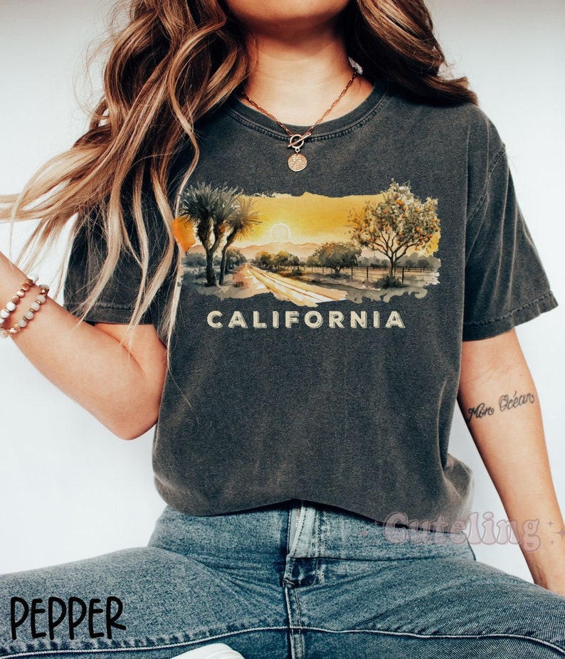 May include: A dark gray t-shirt with a watercolor illustration of a California sunset and the word "CALIFORNIA" printed in yellow. The shirt is worn by a person with a gold necklace and blue jeans.