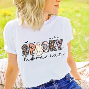 May include: White t-shirt with a colorful "Spooky Librarian" graphic featuring a ghost and bats.