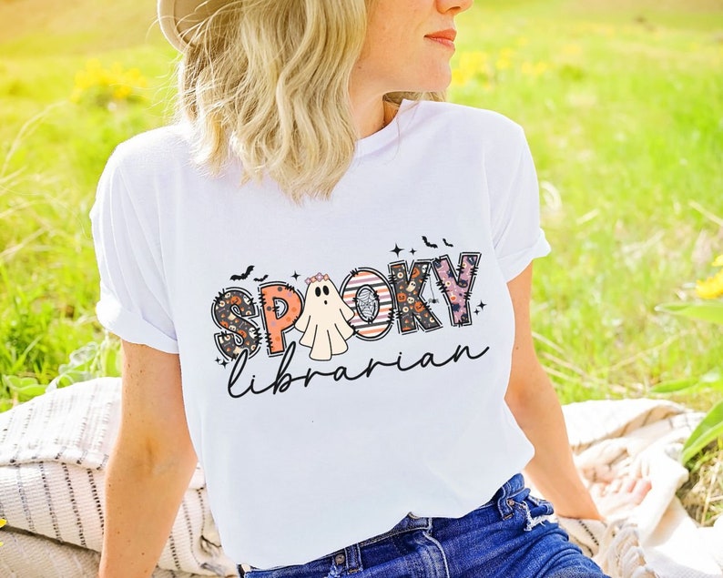 May include: White t-shirt with a colorful "Spooky Librarian" graphic featuring a ghost and bats.