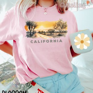 May include: A pink t-shirt with a watercolor illustration of a California landscape with palm trees, a road, and a sunset. The text "CALIFORNIA" is printed below the image.