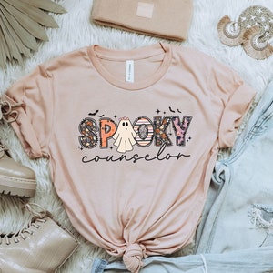 May include: A pink t-shirt with the text "SPOOKY counselor" in colorful letters with a ghost in the middle of the word "SPOOKY".