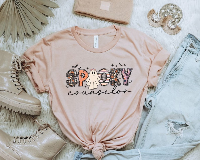 May include: A pink t-shirt with the text "SPOOKY counselor" in colorful letters with a ghost in the middle of the word "SPOOKY".