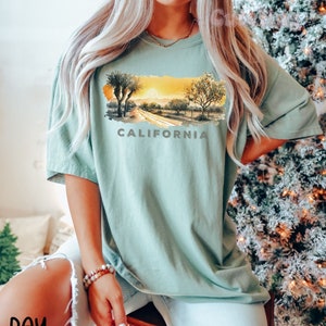 May include: A light green t-shirt with a watercolor graphic of a sunset over a road in California. The text "CALIFORNIA" is printed below the graphic.