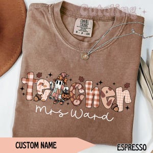 May include: Espresso-coloured t-shirt with the word "teacher" in a fall-themed design, featuring a ghost in a witch hat holding a pumpkin. The name "Mrs. Ward" is written below. A silver necklace is draped across the shirt.
