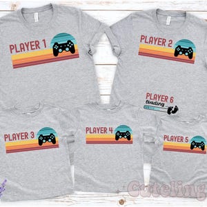 May include: Gray t-shirts with a retro design featuring a game controller and the words "Player 1" through "Player 5". One shirt says "Player 6 loading" with a loading bar and footprints. The design includes a sunset-colored stripe.
