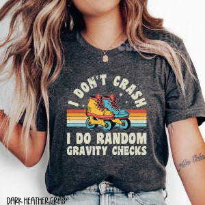Pu&ograve; includere: Maglietta grigio scuro melange con un design arcobaleno retr&ograve; e un paio di pattini a rotelle. Il testo sulla maglietta recita "I don't crash I do random gravity checks".