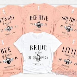 Könnte beinhalten: Fünf pfirsichfarbene T-Shirts mit schwarzem Text und Bienen-Grafiken. Die Shirts zeigen die Aufschriften "Let's Get Buzzed", "Bee Hive", "She Found Her Honey", "Bee of Honor" und "Little Honey Bee".