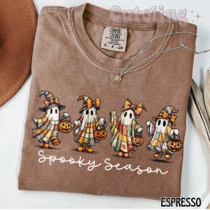 Op de afbeelding: Espresso-kleurig T-shirt met een afbeelding van vier cartoongeesten in Halloween-kostuums, elk met een pompoen-lantaarn. De tekst "Spooky Season" staat eronder. Het shirt heeft een Comfort Colors-label.