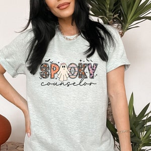 May include: A light gray t-shirt with a graphic print of a ghost and the text "Spooky Counselor" in colorful letters with floral and Halloween-themed accents.