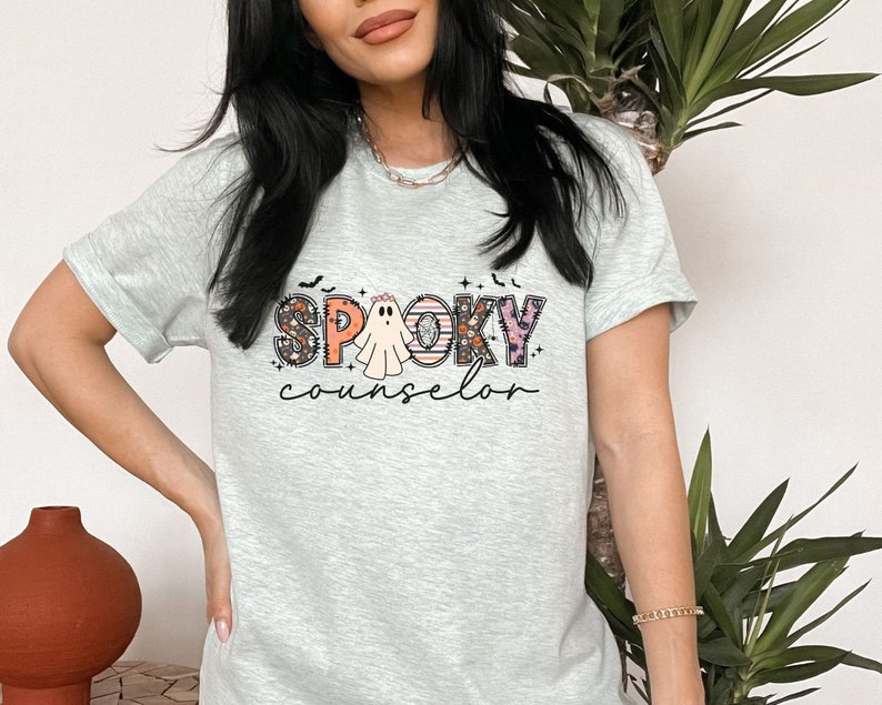 May include: A light gray t-shirt with a graphic print of a ghost and the text "Spooky Counselor" in colorful letters with floral and Halloween-themed accents.