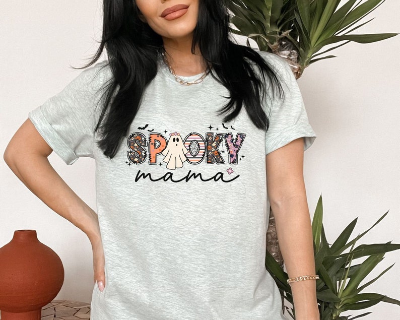 May include: A light gray t-shirt with a graphic print of the words "Spooky mama" in colorful letters with a ghost in the middle. The letters are decorated with floral and Halloween themed patterns.