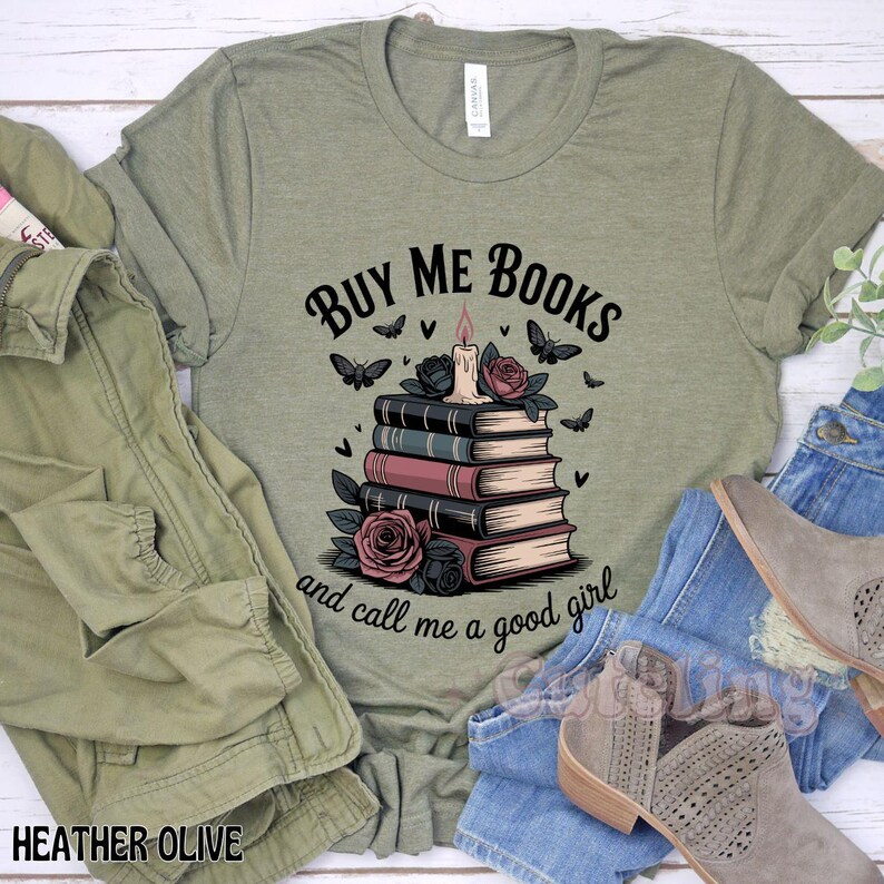 May include: Heather olive green t-shirt with the text "BUY ME BOOKS and call me a good girl." The graphic features a stack of books, a candle, roses, and butterflies. The shirt is surrounded by a green jacket, jeans, and boots.