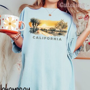 May include: A light blue t-shirt with a watercolor illustration of a California sunset with palm trees and the word "CALIFORNIA" printed in gray below the image. The t-shirt is made of a soft, comfortable fabric and has a relaxed fit.