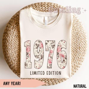 Può includere: T-shirt color crema con l'anno "1976" in grandi numeri a fantasia floreale. Il testo "LIMITED EDITION" è sotto i numeri. La maglietta è su una superficie circolare intrecciata. Sono visibili anche le parole "ANY YEAR!" e "NATURAL".