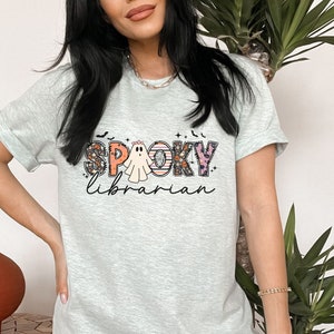 May include: A light gray t-shirt with a floral print design that spells out "Spooky Librarian" with a ghost in the "O".
