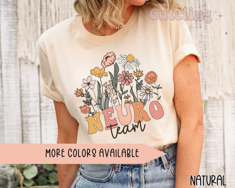 Puede incluir: Una camiseta beige claro con un dise&ntilde;o floral y el texto "Neuro Team" en rosa, amarillo y naranja. El texto "M&aacute;s colores disponibles" est&aacute; impreso debajo del dise&ntilde;o. La camiseta la lleva puesta una persona con el pelo largo y rubio.