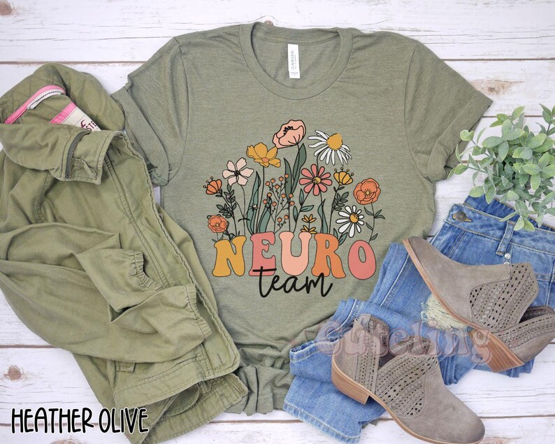 Puede incluir: Una camiseta verde con un dise&ntilde;o floral y el texto "Neuro team" en letras de colores. La camiseta est&aacute; sobre una superficie de madera con un par de vaqueros y botas.