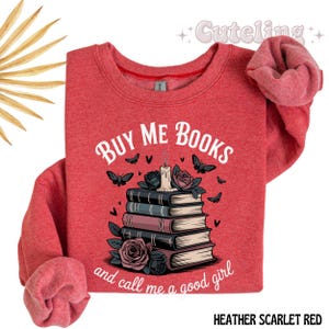 May include: Heather scarlet red sweatshirt with the text "BUY ME BOOKS and call me a good girl". The graphic features a stack of books, roses, butterflies, and a candle.