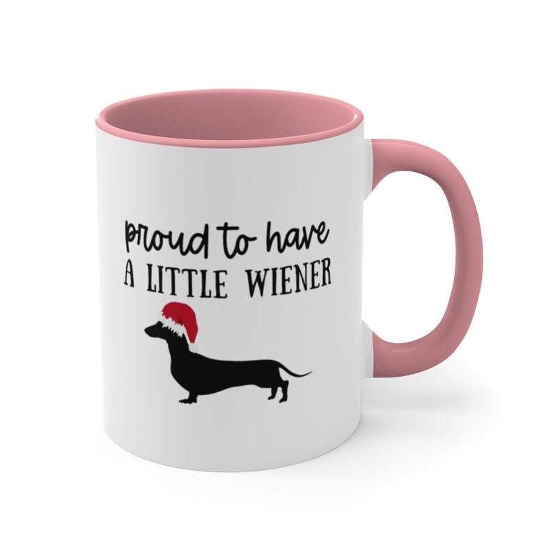 May include: White ceramic mug with a pink interior and handle. The mug features the text "proud to have a LITTLE WIENER" above a black dachshund wearing a Santa hat. A fun gift for dog lovers.