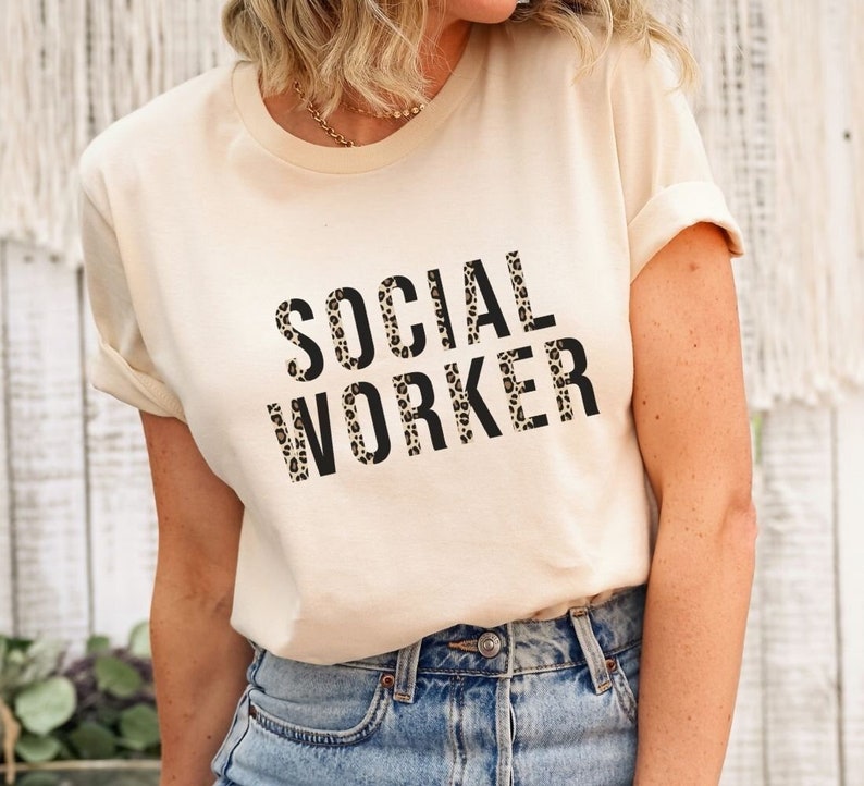 May include: A cream colored t-shirt with the words "SOCIAL WORKER" printed in black letters with a leopard print pattern.