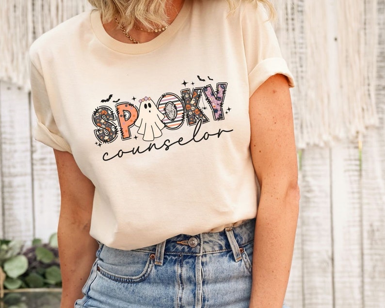 May include: A cream colored t-shirt with a graphic design of the word "SPOOKY" in colorful patchwork letters with a ghost in the middle and the word "counselor" below.