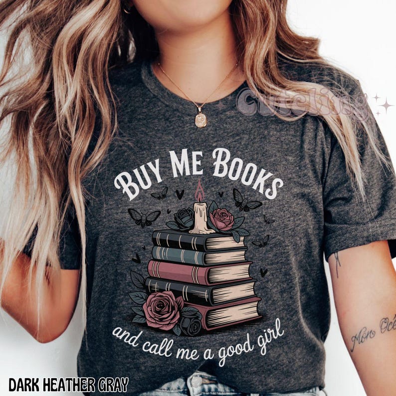 May include: Dark heather gray t-shirt with the text "BUY ME BOOKS and call me a good girl." The graphic features a stack of books, roses, butterflies, and a candle. The words "DARK HEATHER GRAY" are at the bottom.