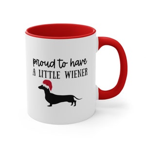 May include: White ceramic mug with a red interior and handle. The mug features the text "proud to have a LITTLE WIENER" above a black dachshund wearing a Santa hat. A fun, festive gift.