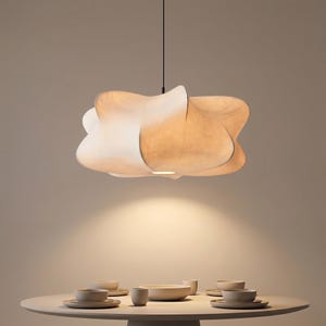 Puede incluir: Una lámpara colgante blanca con forma de nube y textura suave y translúcida cuelga de un cable negro fino sobre una mesa de comedor redonda. La mesa está dispuesta con varios cuencos, platos y tazas pequeñas de cerámica mate clara.