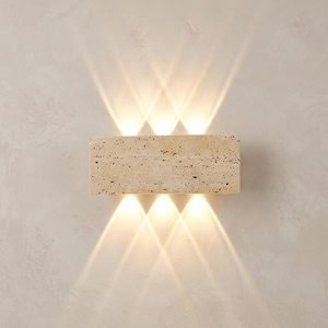 May include: A rectangular wall sconce made of beige, porous travertine stone. The fixture emits three distinct beams of warm, bright light directed upward and three identical beams of light directed downward, creating a symmetrical lighting effect.