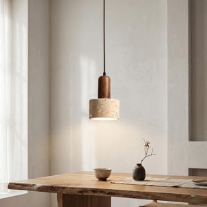 May include: A pendant light fixture featuring a cylindrical dark brown wooden top piece connected to a wider, porous beige travertine stone shade. The light hangs from a thin black cord and emits a warm downward glow from the stone cylinder.