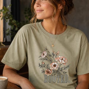 May include: A sage green short-sleeved t-shirt featuring a graphic print of a floral bouquet with pink and white flowers and green leaves. Below the floral illustration, the word "ROOTED." is printed in a bold, dark gray serif font.