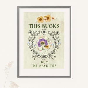Może przedstawiać: Oprawiony wydruk ścienny na kremowym tle z napisem THIS SUCKS BUT WE HAVE TEA ciemnozielonymi literami. Projekt zawiera centralnie umieszczony fioletowy kwiat, żółte akcenty kwiatowe oraz czarny wieniec botaniczny wykonany kreską.