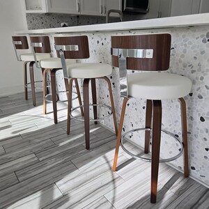 Könnte beinhalten: Ein Set aus vier drehbaren Barhockern mit geschwungenen Rückenlehnen aus dunklem Holz, weißen, gepolsterten runden Sitzen und verchromten Metallgestellen mit integrierten Fußstützen. Jeder Hocker hat eine T-förmige Chromstütze am Rücken.